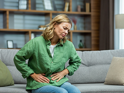 Unhealthy senior woman sitting on sofa at home. holding her stomach, grimacing in pain. She experiences severe abdominal pain, eating disorder, diarrhea, poisoning, menstruation, menopause.
