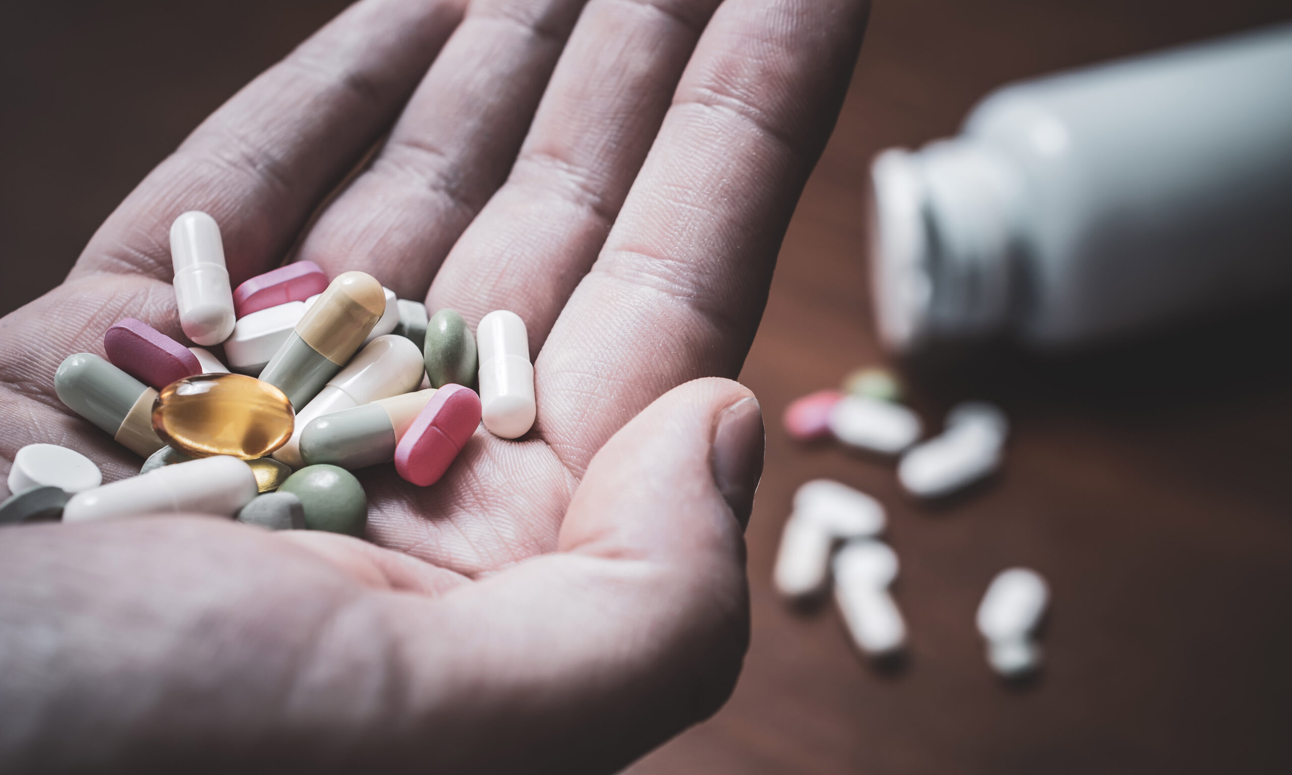 A Macro Of A Male Hand Holding Several Pills With Prescription Bottle And More Pills In Background - Taking Too Many Pills Concept