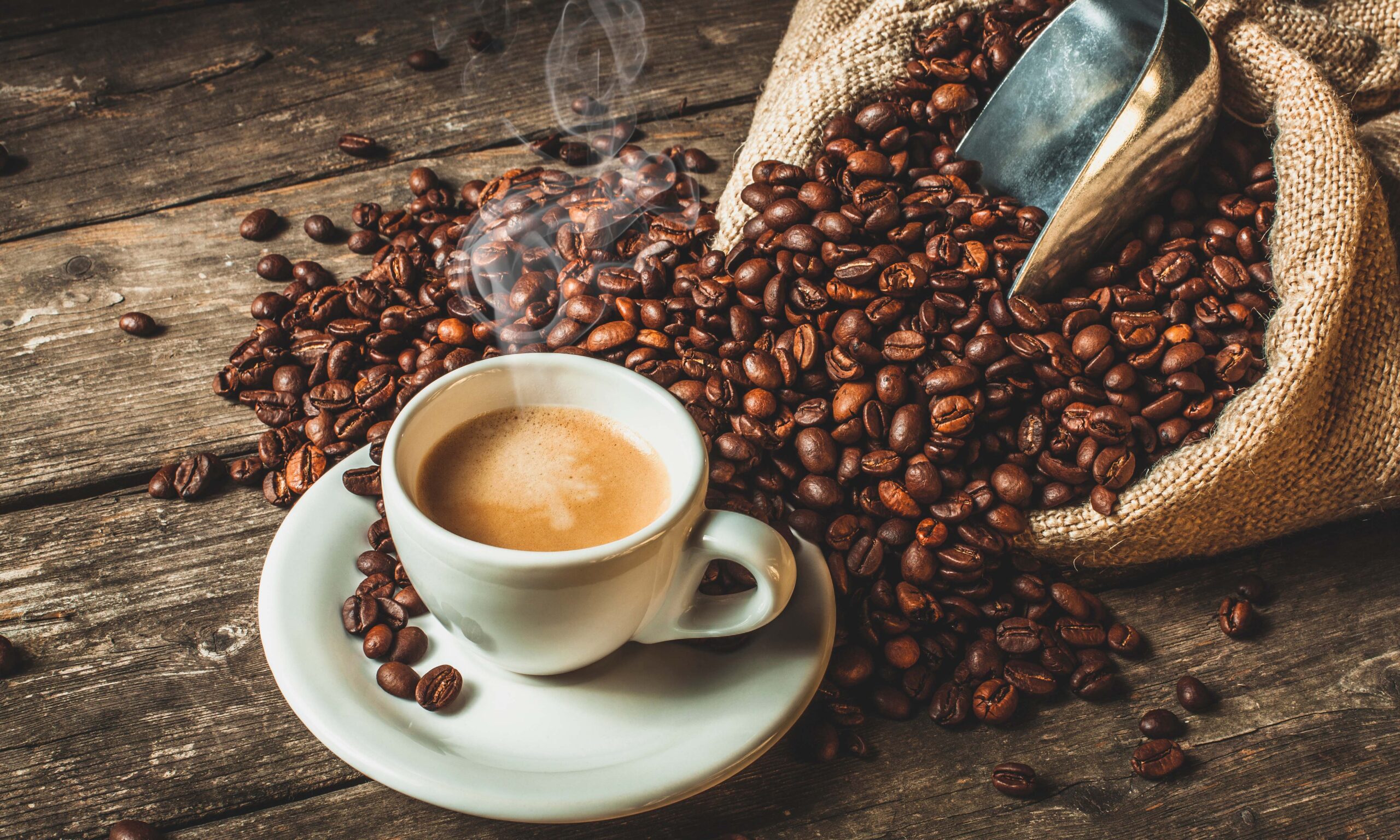 steaming coffee in traditional ceramic cup, jute sack with roasted beans on wooden table.