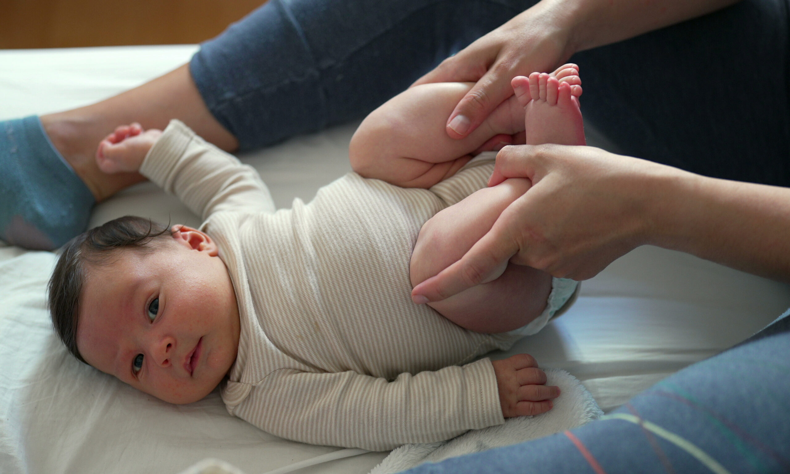 Mother holding baby's feet and gently bicycling his legs to help him relieve gas. Bright and cheerful atmosphere with the baby looking curiously. Cozy home setting