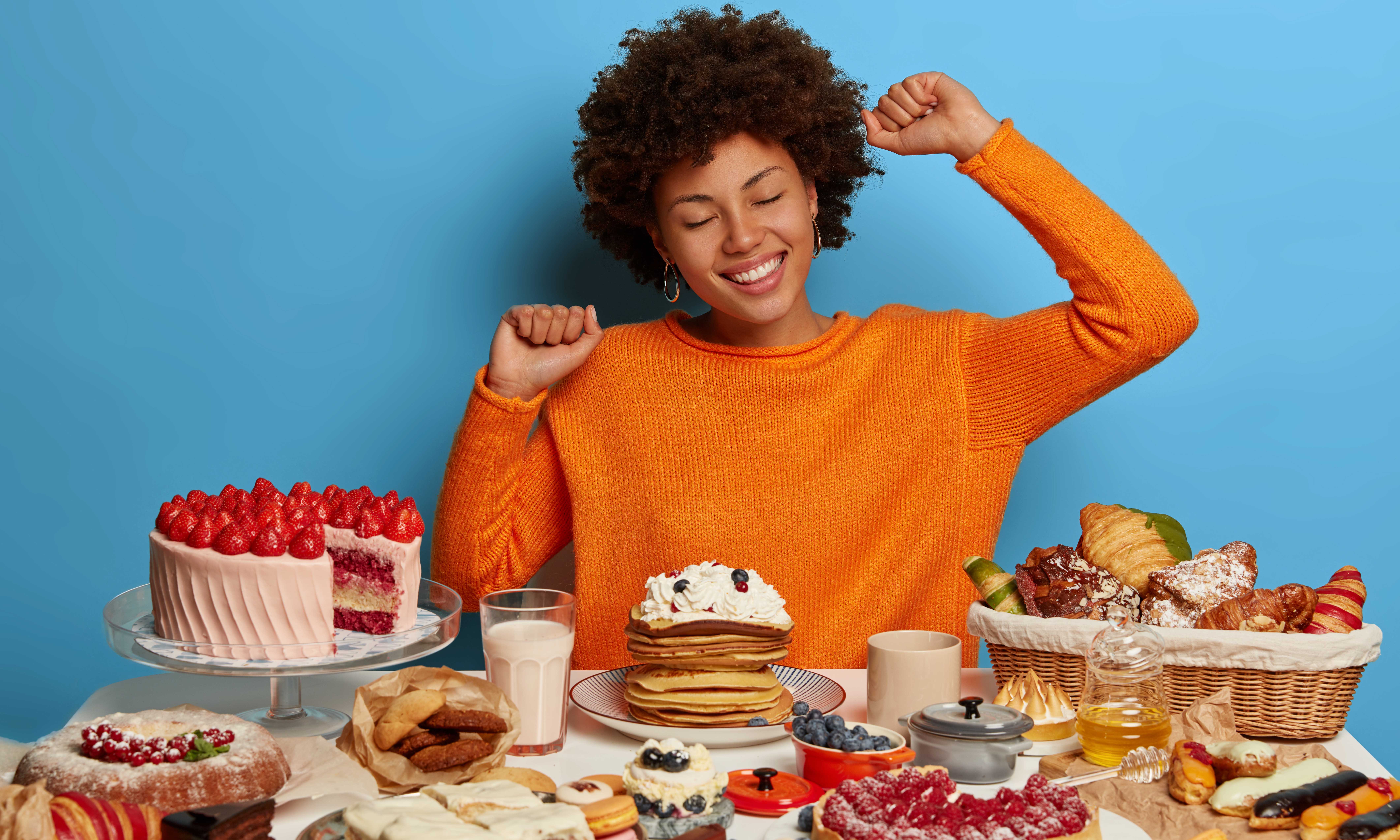 Sweet tooth, temptation by delicious food. Pretty woman with Afro hairstyle has good mood after eating tasty delicacy, stretches at table with confectionery. Laziness, overeating and gluttony