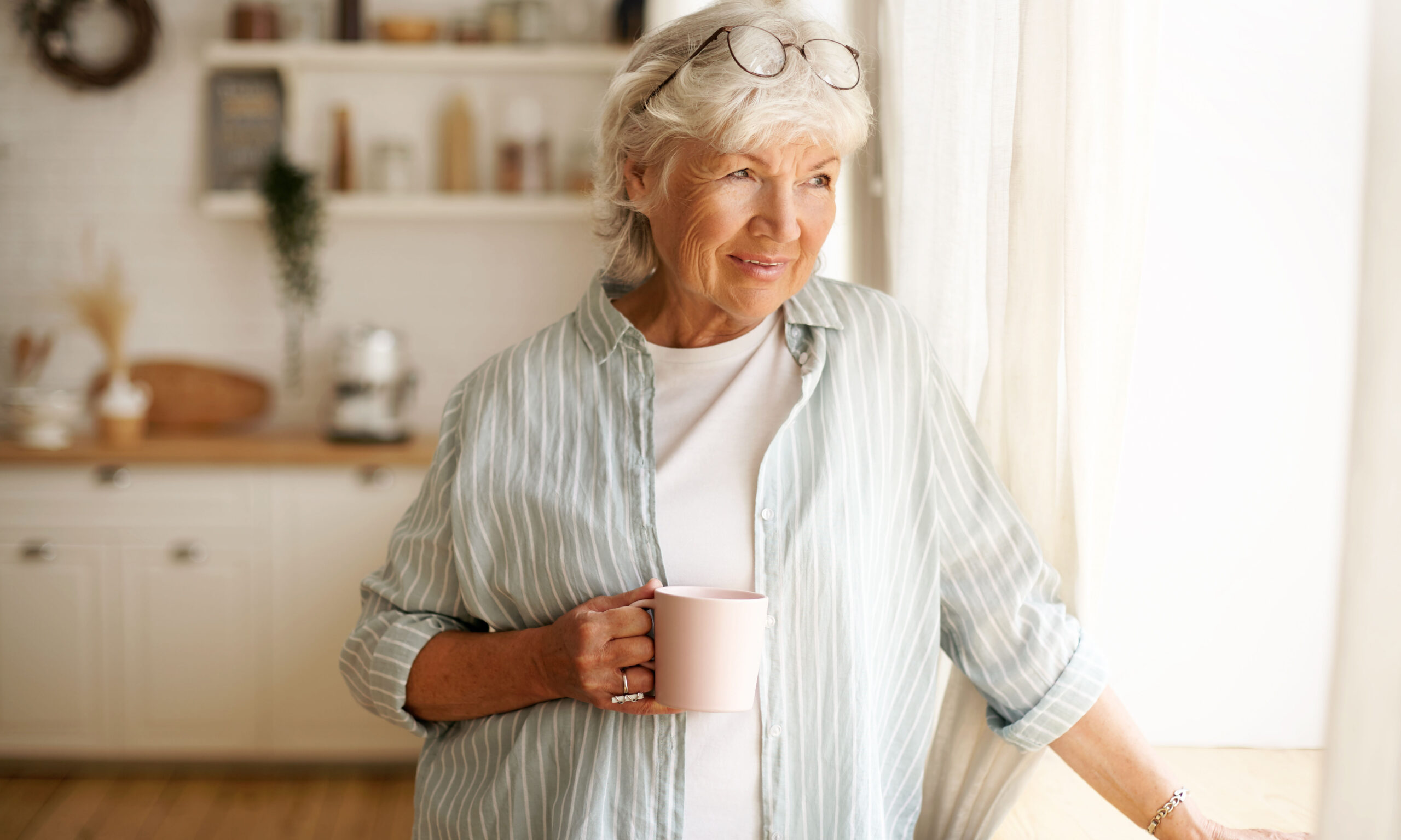 Coziness, domesticity and leisure concept. Portrait of stylish gray haired woman with round spectacles on her head enjoying morning coffee, holding mug, looking outside through window glass