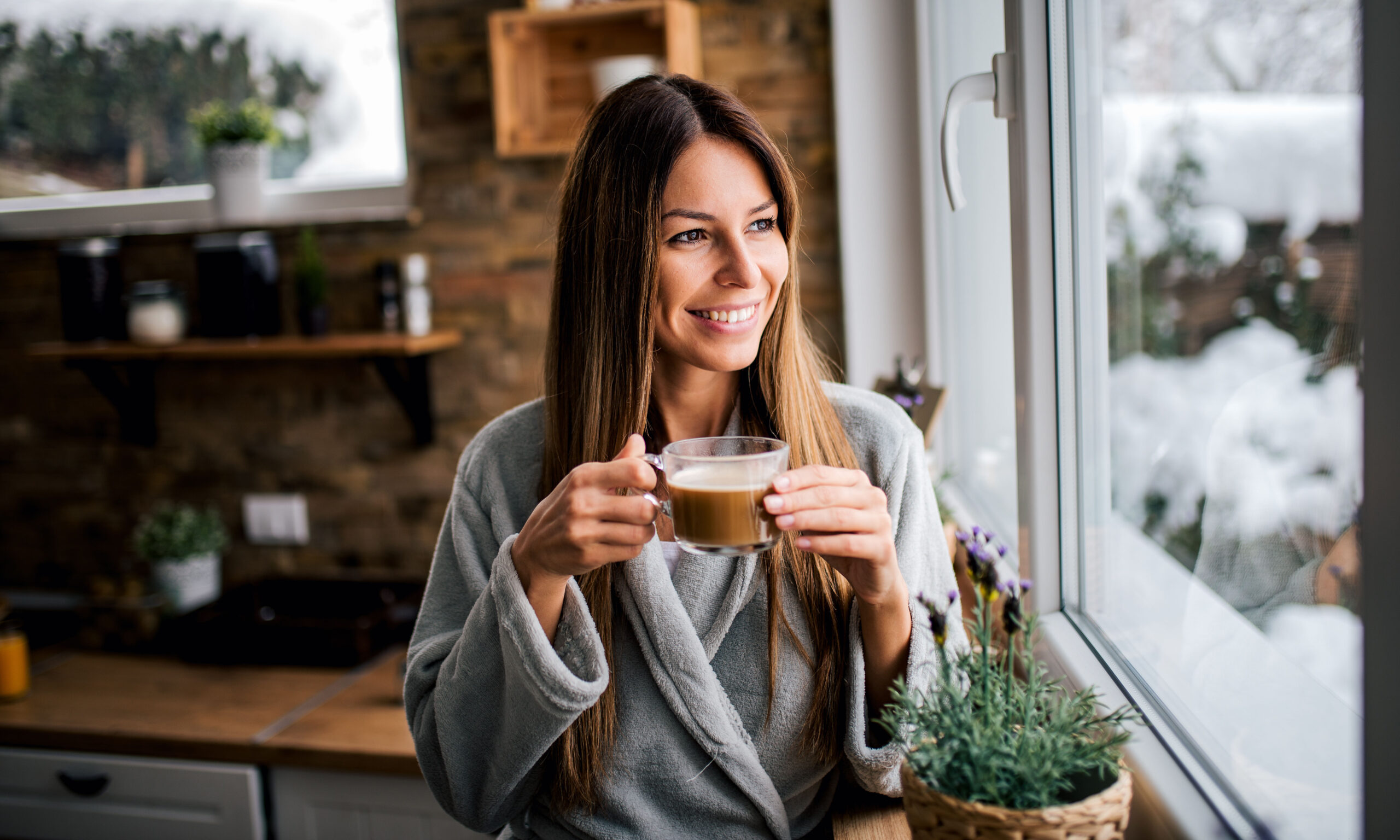 Beautiful smiling brunette looking through window and drinking coffee in the kitchen.