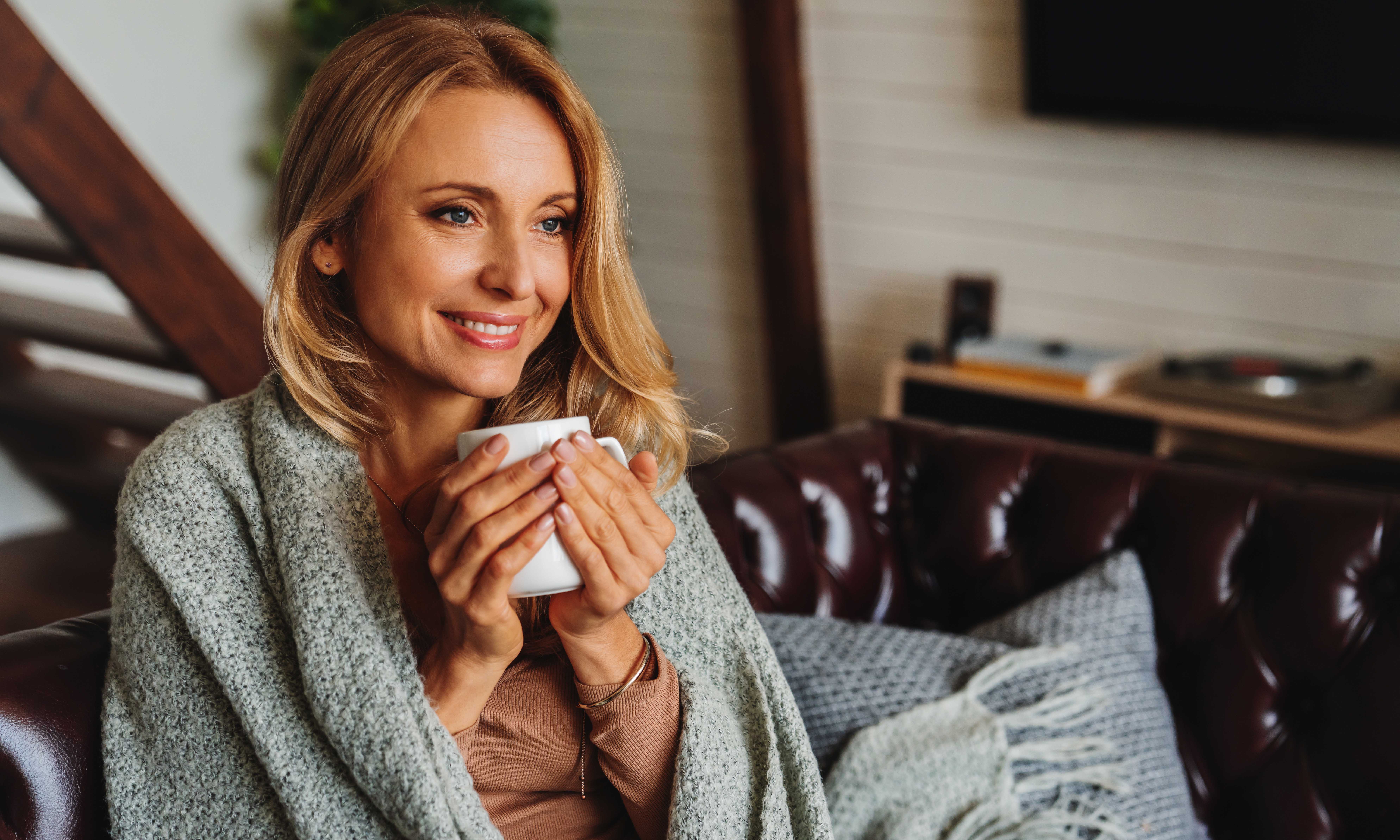 Close up of dreaming middle aged woman sitting in living room with cup of coffee or tea enjoying under blanket