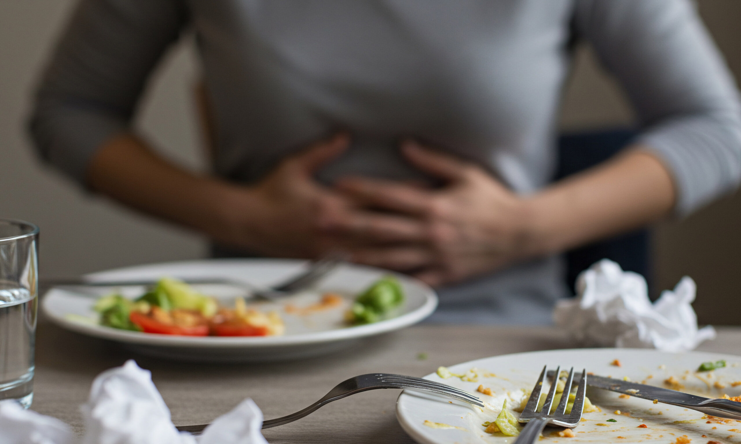 Blurred woman holds stomach in pain after eating, focus on dirty plates, cutlery, crumpled napkins, and glass of water.