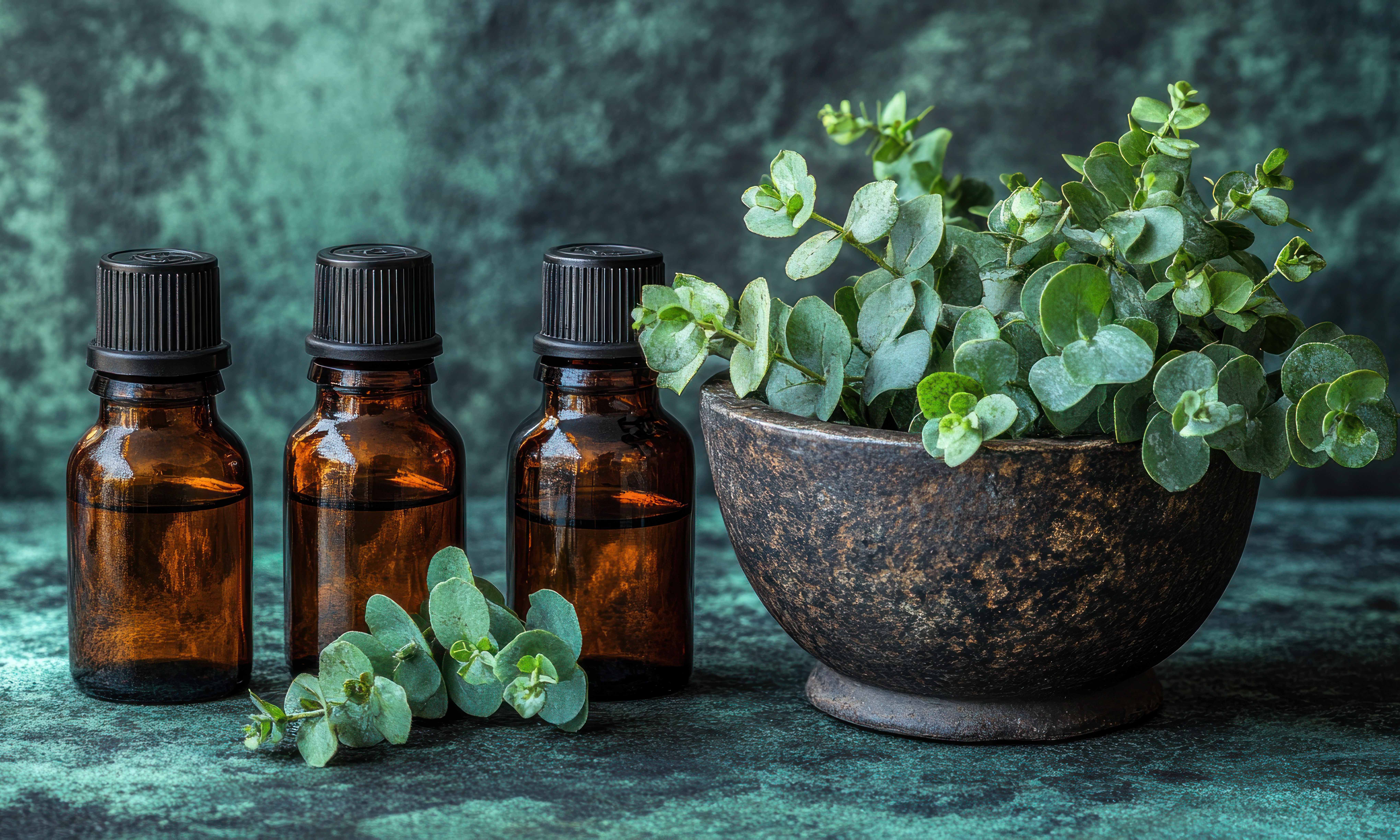 Three amber glass bottles of essential oil, alongside fresh eucalyptus leaves in a dark bowl, set against a teal-green textured backdrop