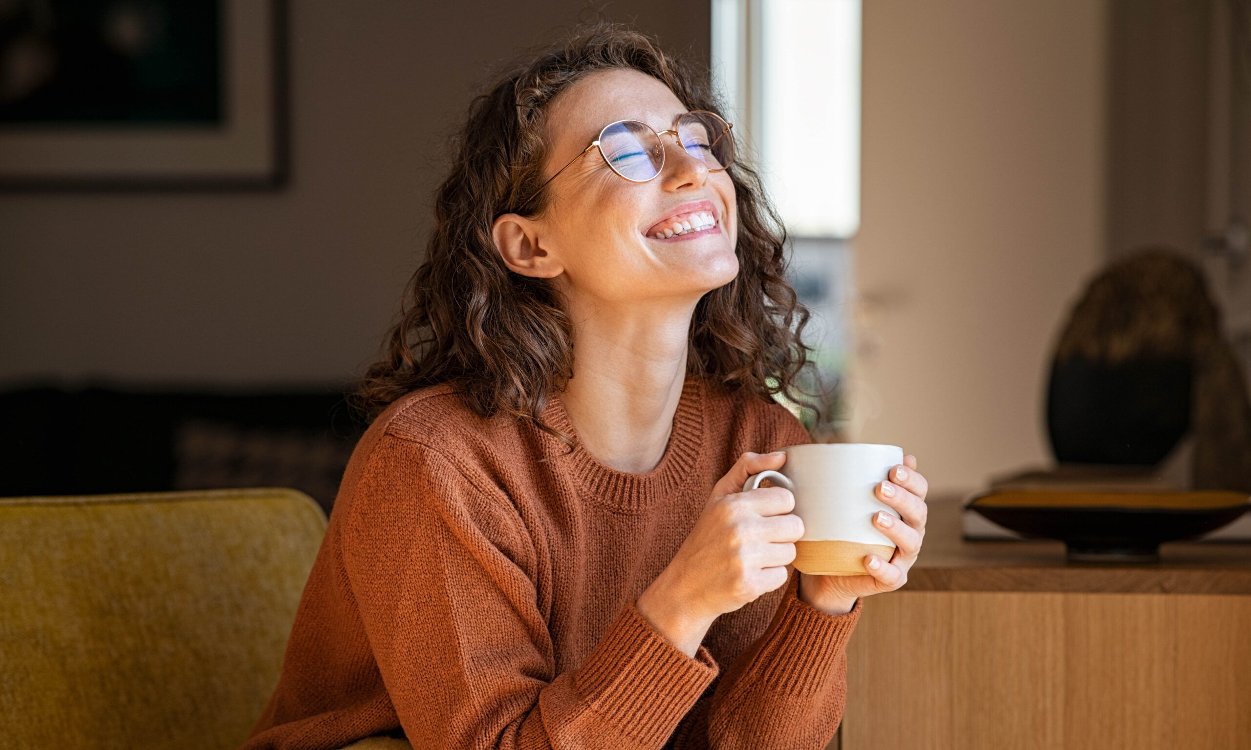 Portrait of joyful young woman enjoying a cup of coffee at home. Smiling pretty girl drinking hot tea in front of the window in winter. Excited woman wearing spectacles and sweater and laughing in an autumn day.