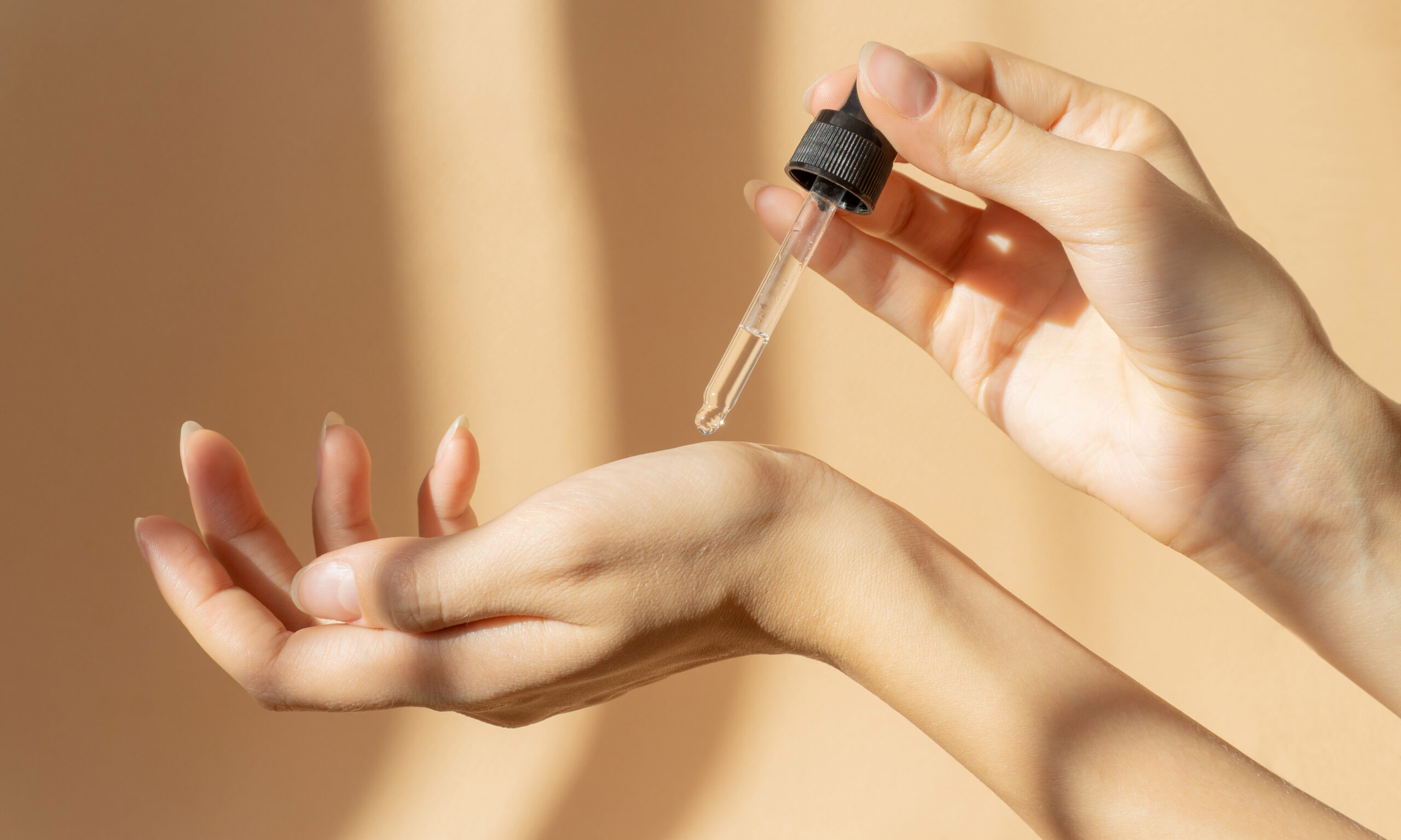 Women's hands hold pipette with cosmetic serum on light brown background, in rays of sunlight. Close-up, copy space. Concept of body care.