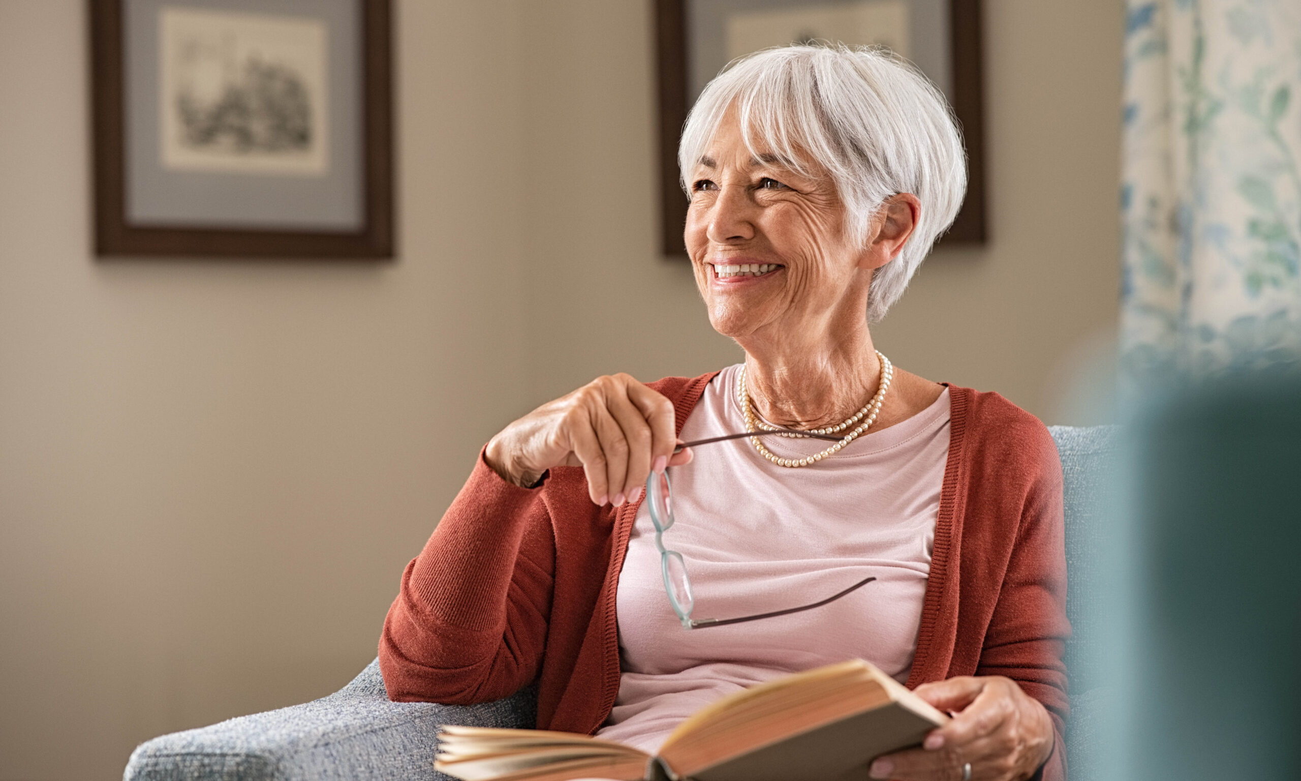 Cheerful senior woman holding book and eyeglasses thinking while relaxing at home. Happy elderly woman reading book at home sitting on couch. Beautiful old teacher takes a break from reading while looking through the window with a big grin.
