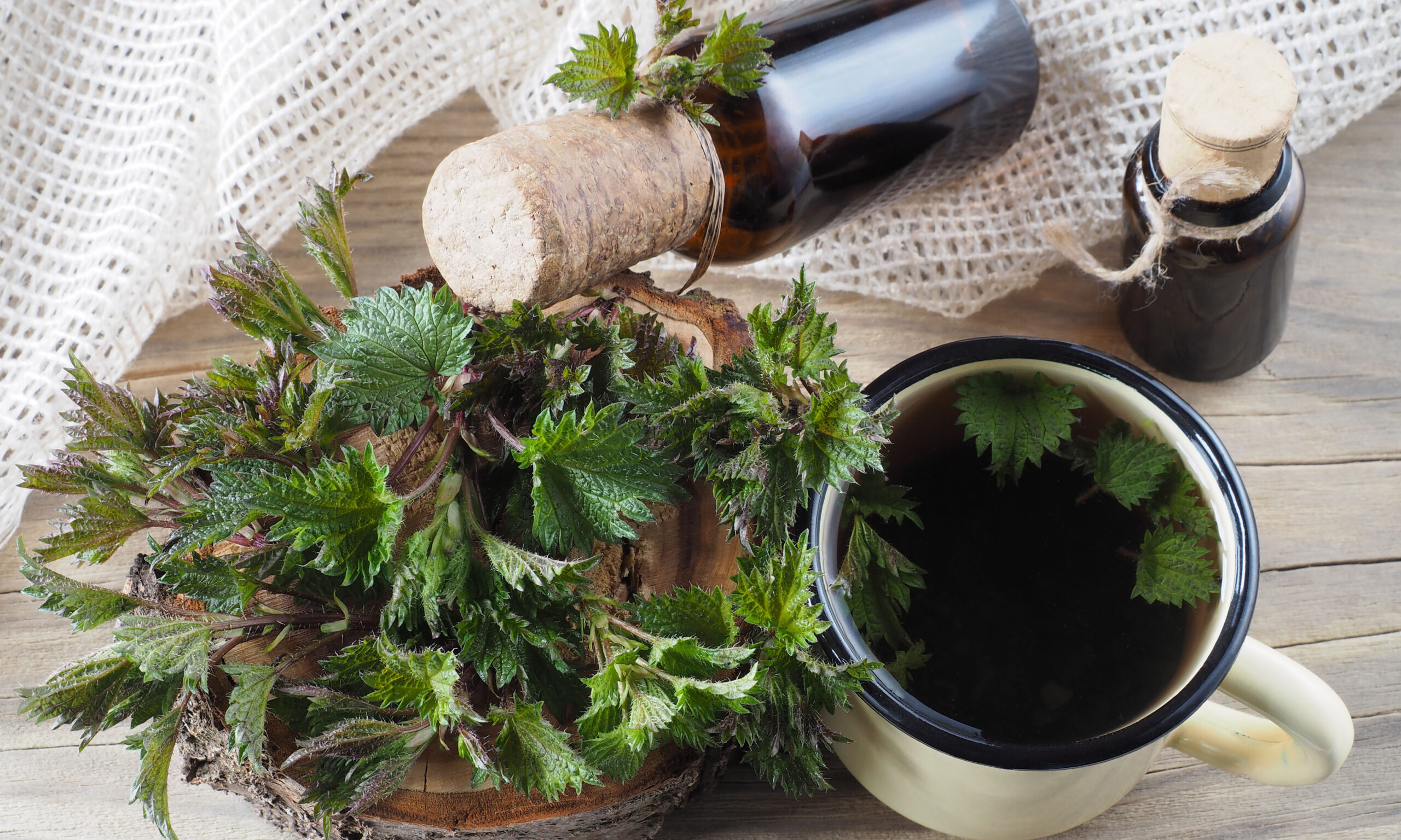 Fresh young nettle leaves, herbal drink in a mug, juice, tincture in a bottles on a wooden background, top view. Medicinal plants urtica for use in food, medicine, homeopathy and cosmetology