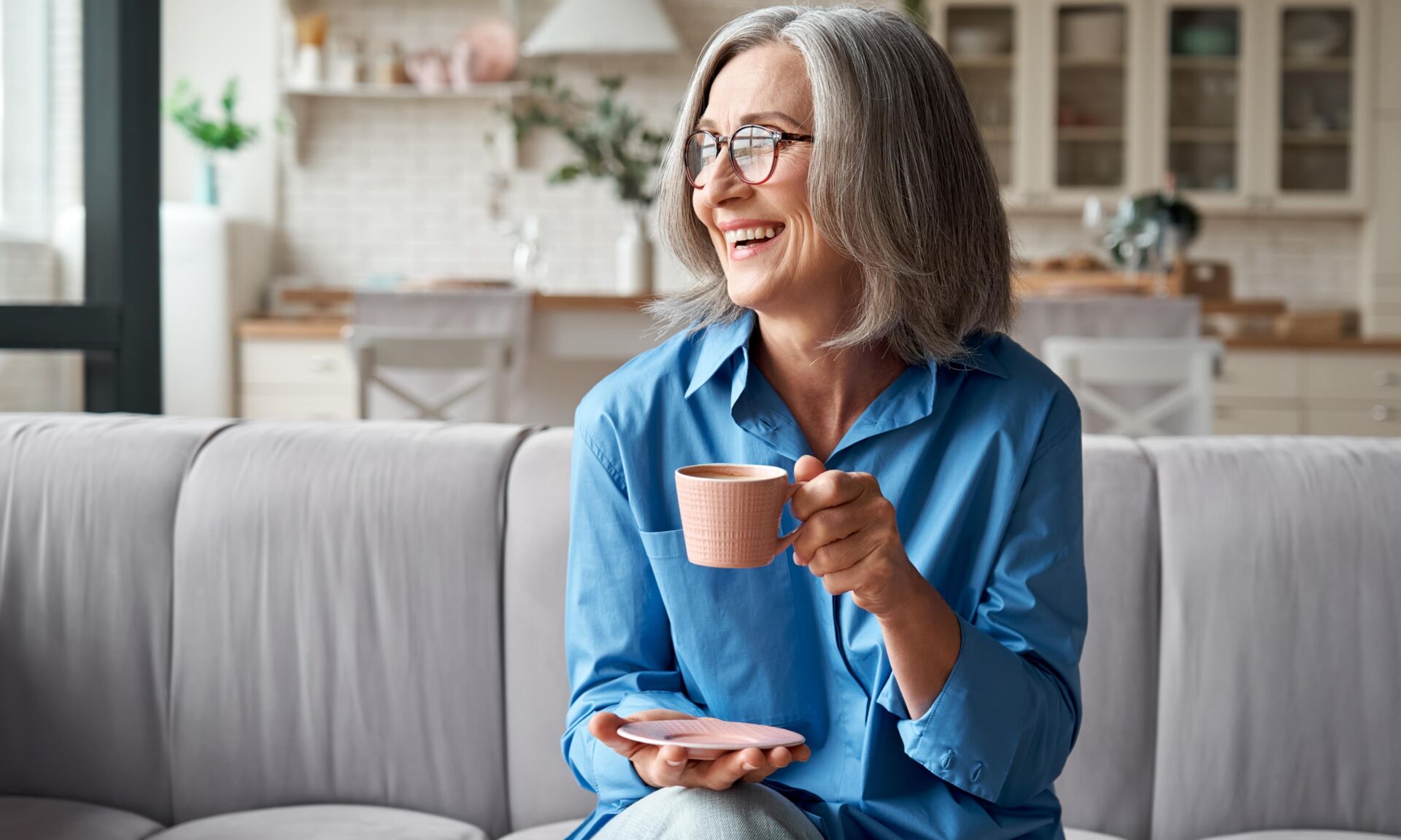 Happy beautiful relaxed mature older adult grey-haired woman drinking coffee relaxing on sofa at home. Smiling stylish middle aged 60s lady enjoying resting sitting on couch in modern living room.