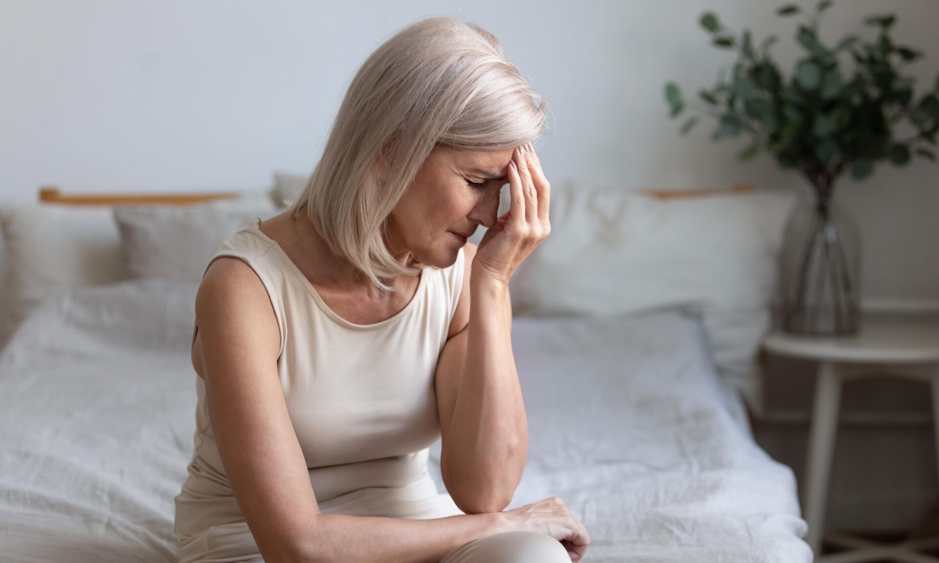 Sick older mature woman sit on bed feel pain dizziness coping with morning headache concept, upset tired middle aged adult woman touching sore head suffer from terrible migraine mental problem