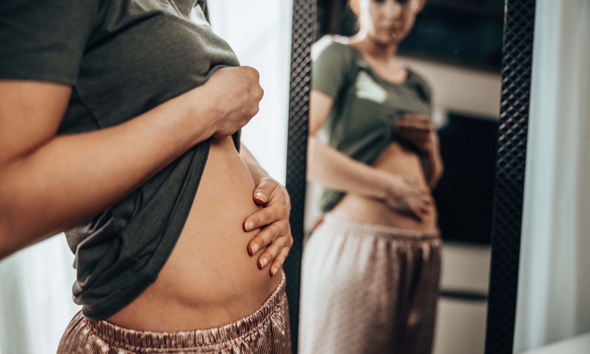 Unhappy young woman standing in front of a mirror and holding hands on her bloating stomach.