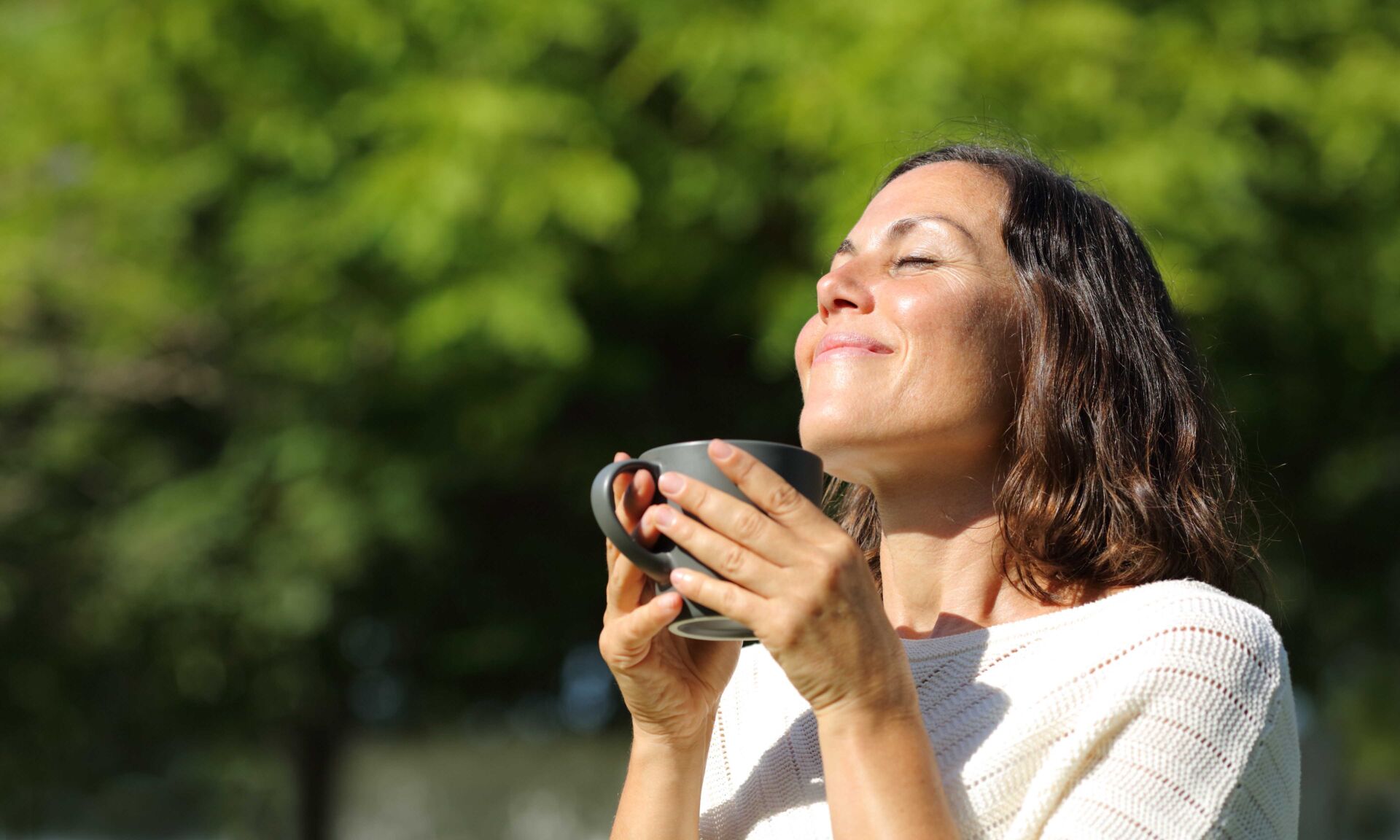Satisfied adult woman smelling coffee cup in a park