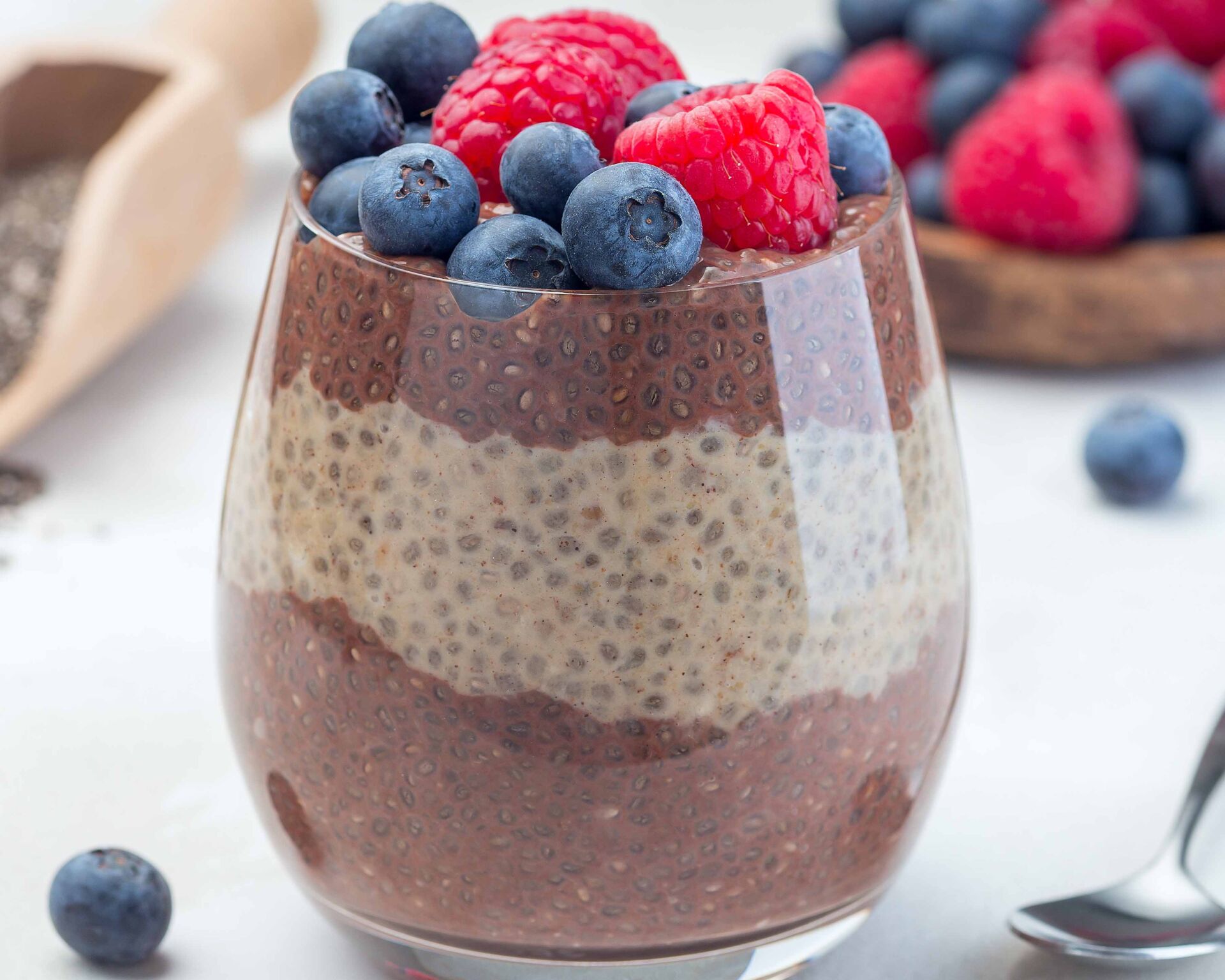 Layered chocolate and peanut butter chia seed pudding in glass, garnished with raspberry and blueberry, square format